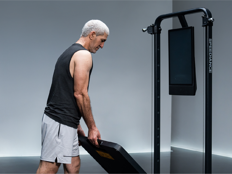 A silver-haired man carrying a training bench towards a Gym Monster
