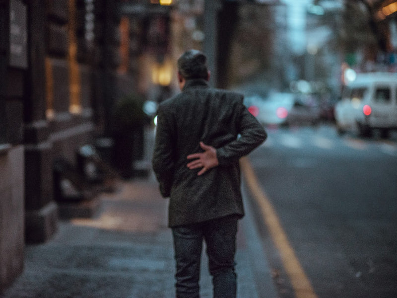 A man in a dark coat on a city street, one hand pressed to his lower back